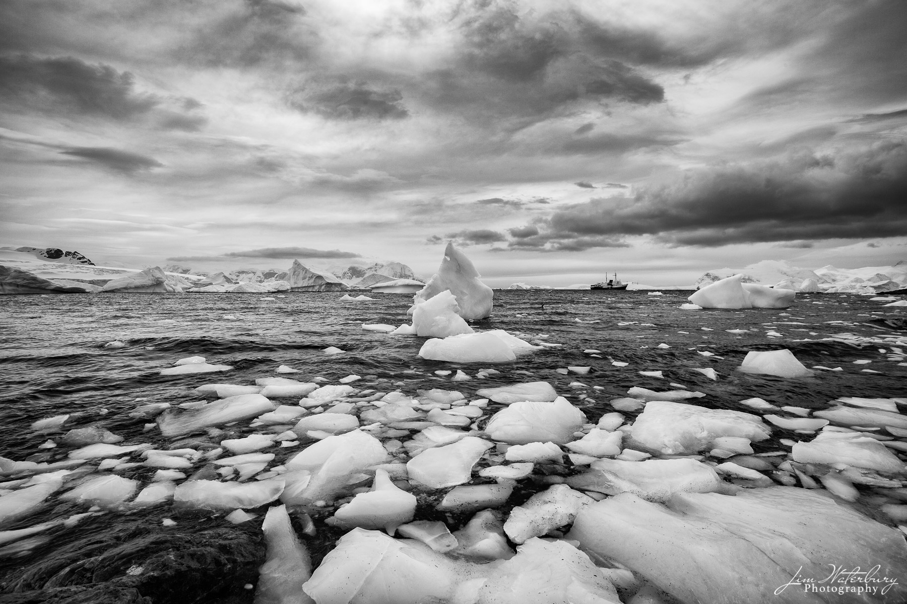Stepping Stones Black & white image of broken ice in a bay off Cuverville Island under stormy skies, with the ship SS Ushuaia in the distance...