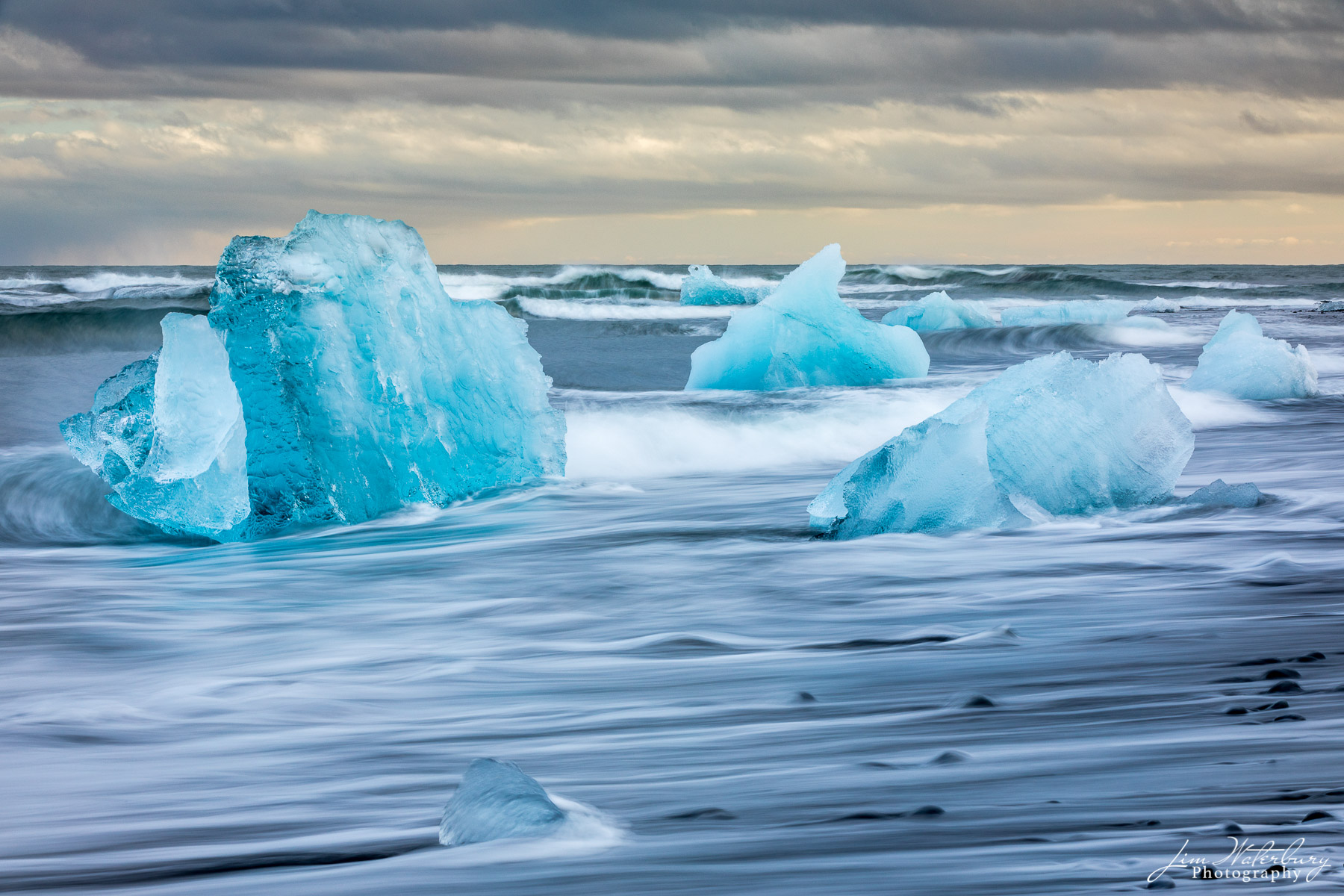 Battered Small icebergs, with their vibrant blue hue, are anchored on the black sand beach of Fellsfjara, and battered by an endless stream...