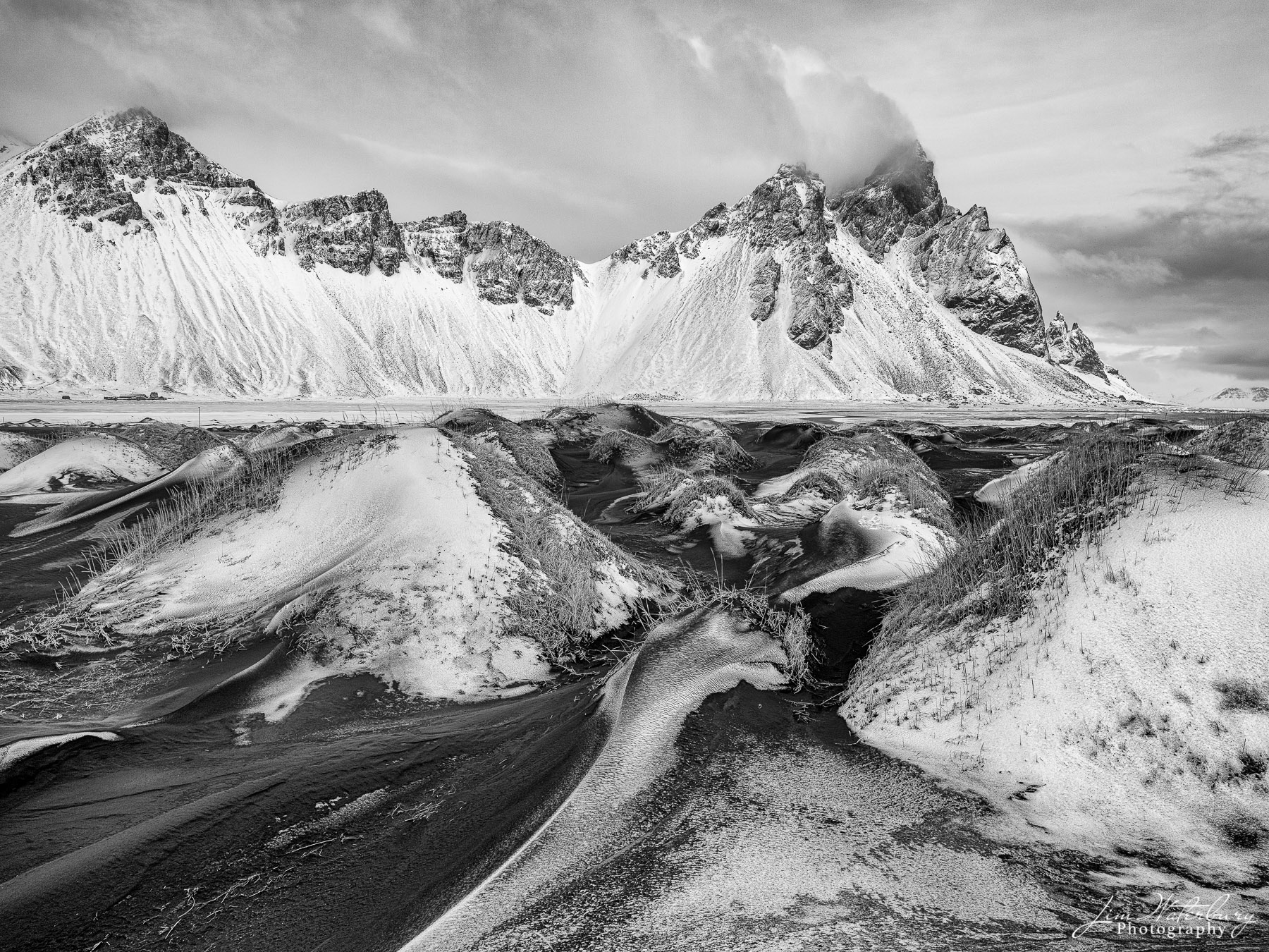 Black & white image of the mounds of black sand at Stokksnes beach in Iceland, covered by a dusting of snow, and against the...