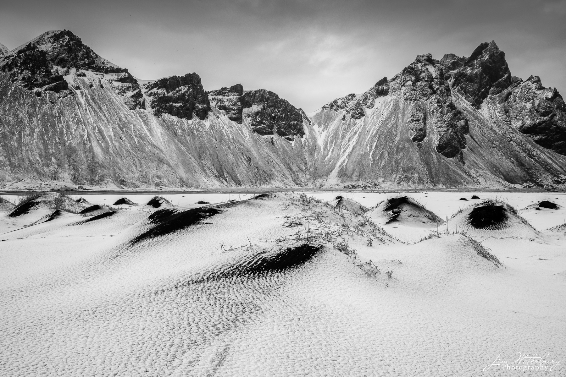 Black & white image of mounds of snow-covered black sand on Stokksnes beach in Iceland resemble small peaks that are mirrored...