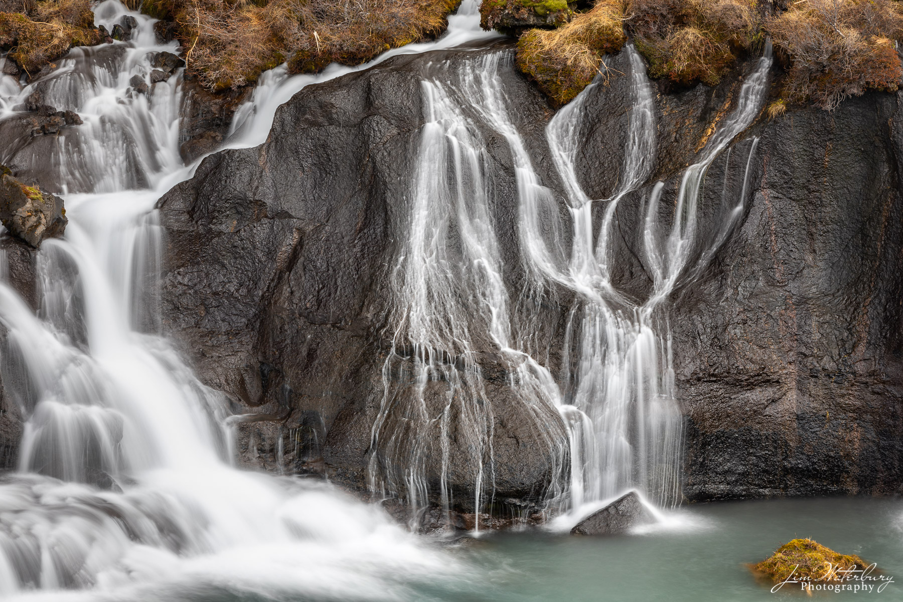 Glacial melt water flows through the lava at the Hraunfossar waterfall in Iceland, forming graceful white ribbons of water against...