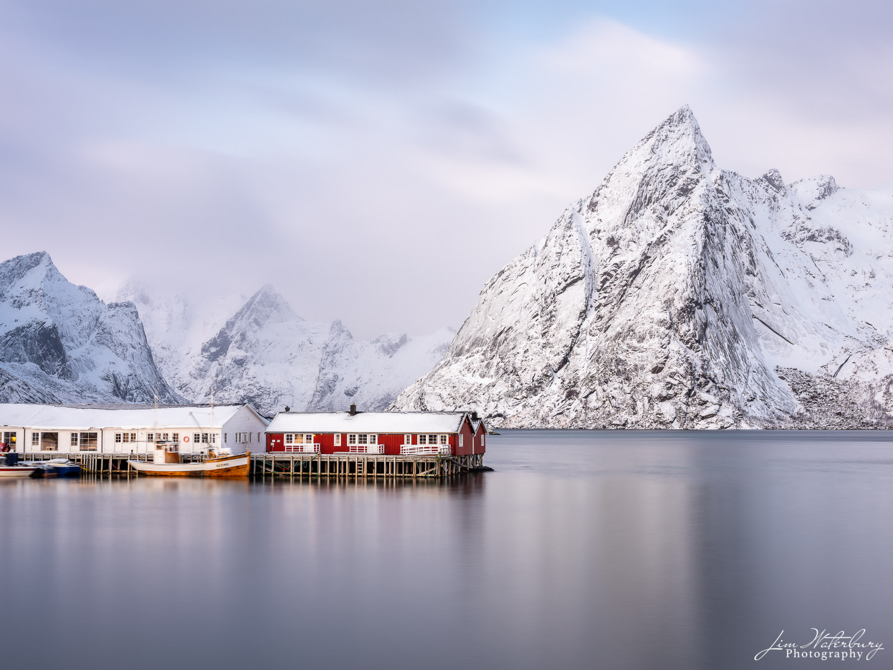 Winter Calm Red fishing cottages (rorbuer) against a snowy mountain backdrop in Hamnoy Harbor, Lofoten, Norway, in winter.