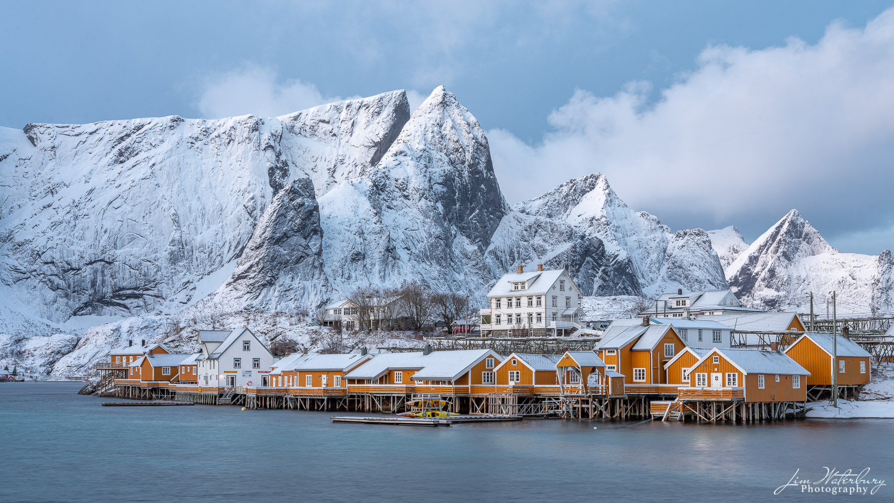 View of the yellow fishing cottages of Sakirsoy, Lofoten, Norway in winter, against a snowy mountain backdrop.