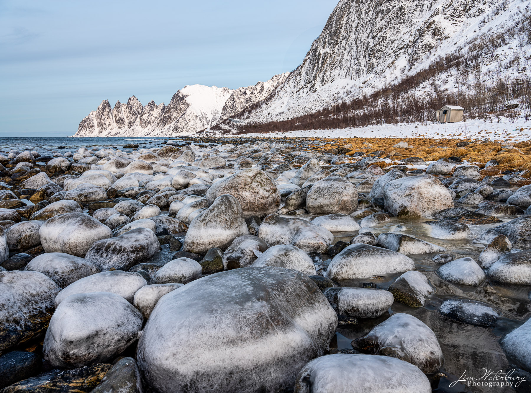 Slippery grass and ice-covered rocks along the shore near Ersfjord, Senja, Norway, against a background of snow-covered mountains...