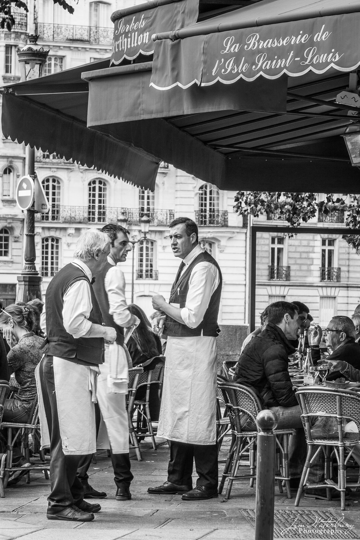 Three Waiters Waiters talk on the sidewalk in front of the Brasserie de l'Ile Saint-Louis, Paris. Black & white.