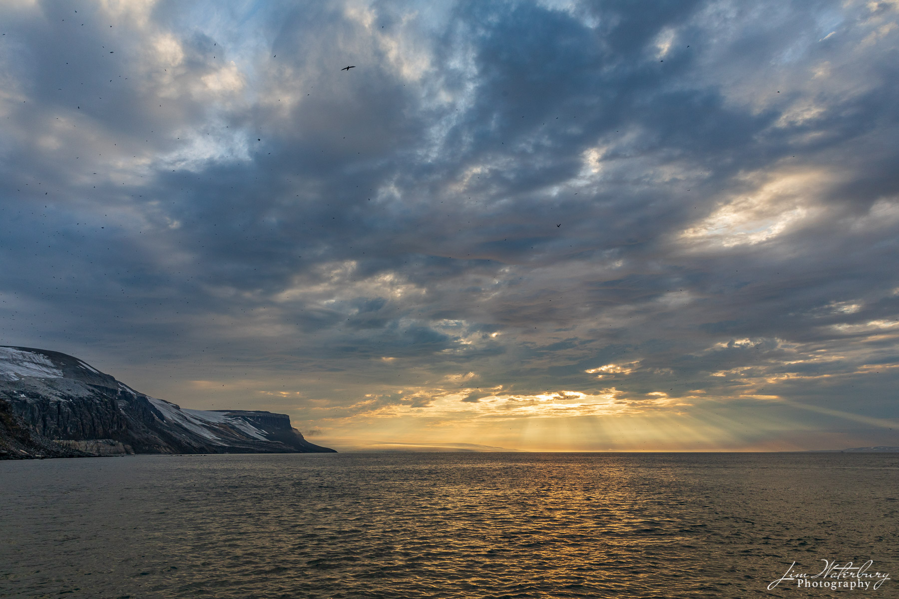 Sunburst Sun, hidden by clouds, casts its rays on the strait near the Alkefjellet bird cliff, Svalbard, Norway. The Hinlopen Strait...